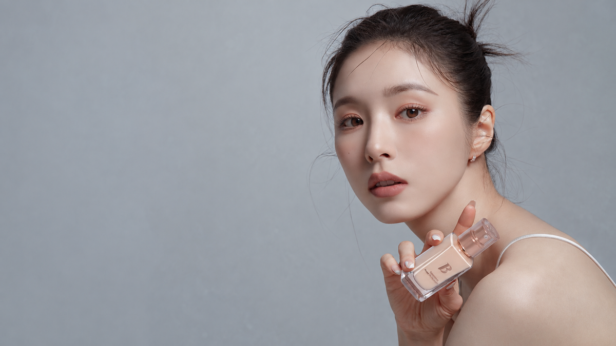 Wide studio portrait of a woman holding a complexion product beside her face, with fresh natural-looking skin and soft gray negative space.