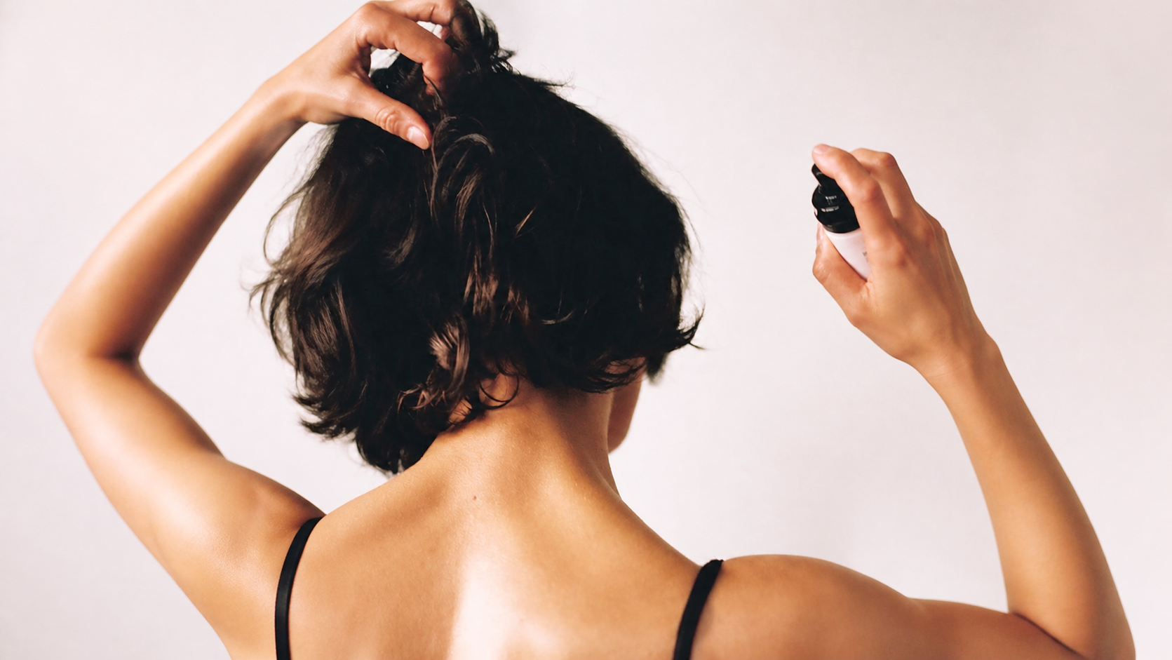Woman viewed from behind lifting dark wavy hair and aiming an unbranded hair-perfume mist toward the mid-lengths against a warm neutral background.