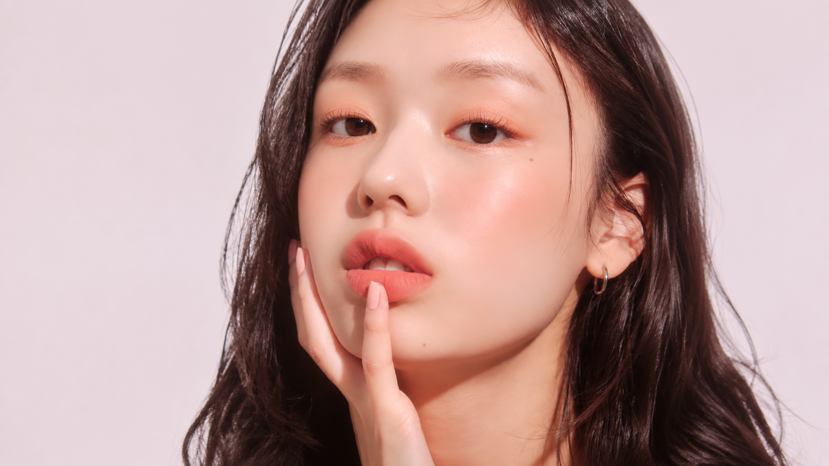 Close-up portrait of a woman wearing soft peach blush and coral makeup against a pale pink background.