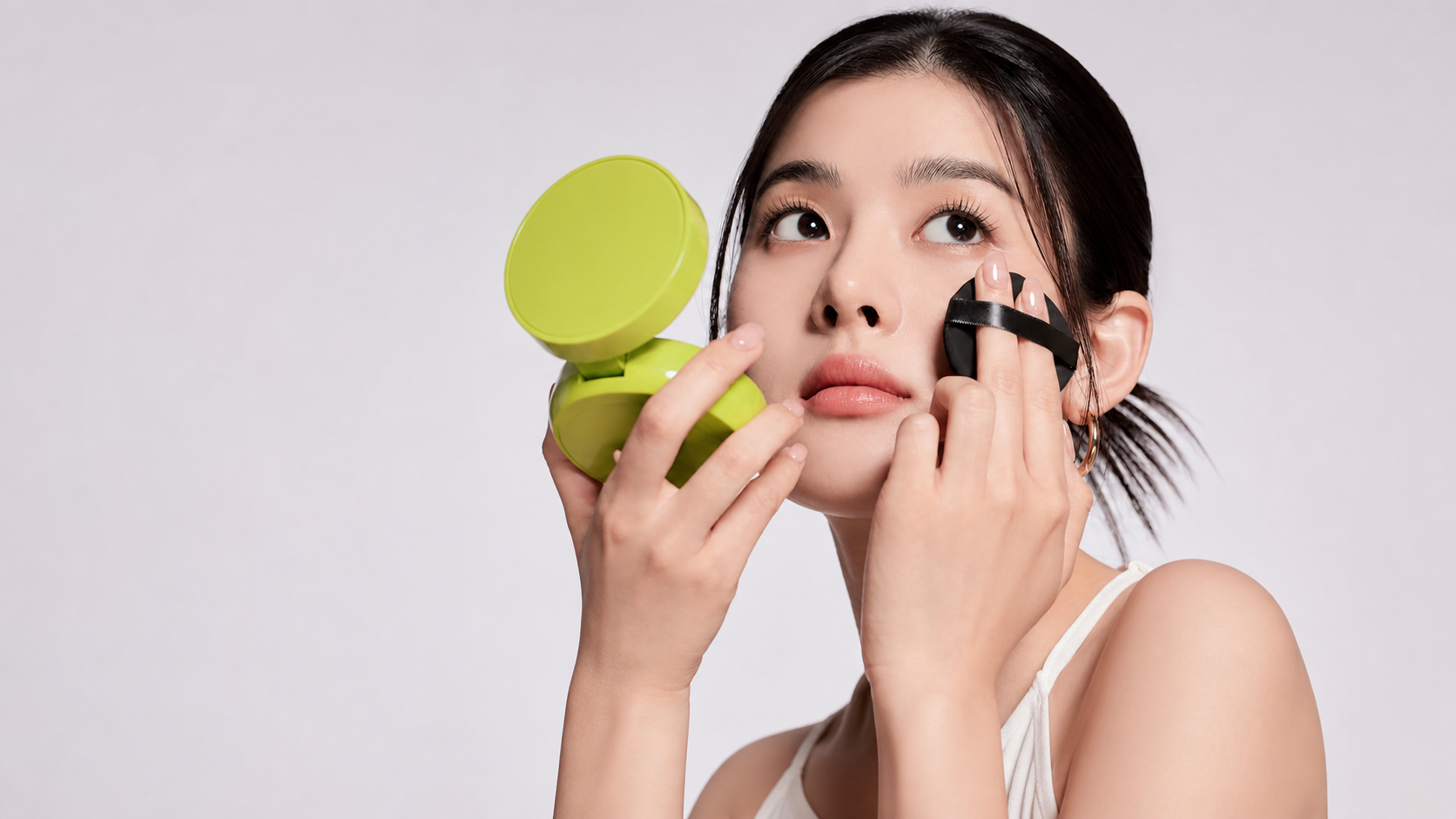 Woman pressing a black cushion foundation puff onto her cheek while looking into a lime compact.