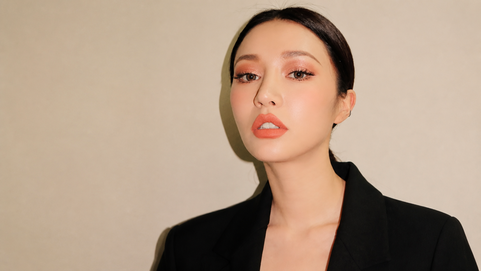 Woman wearing fresh coral eye makeup and fine eyeliner against a warm beige background.