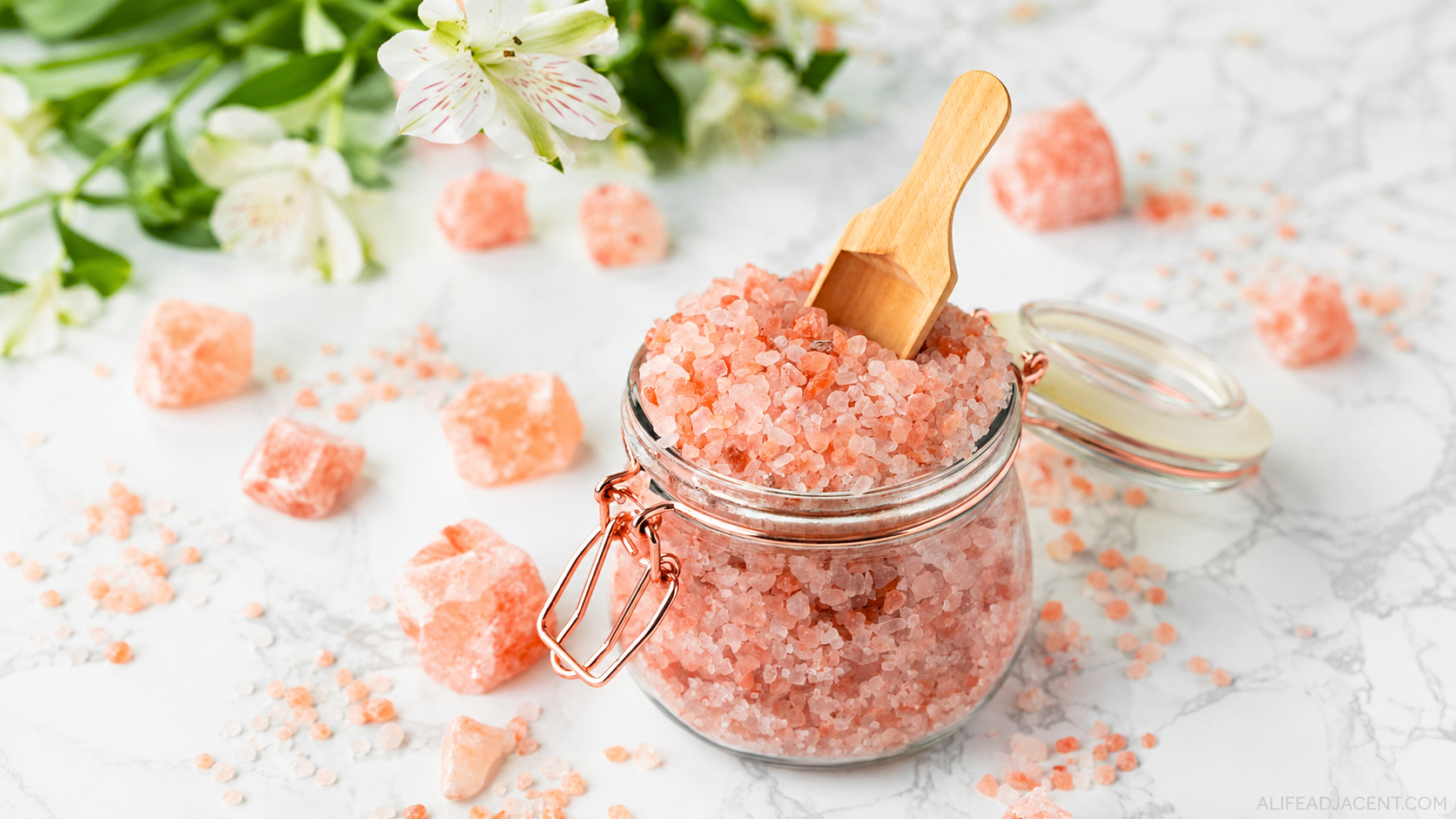 Wide still life of coarse pink mineral salt in a glass jar with a wooden scoop, flowers and scattered crystals on white marble
