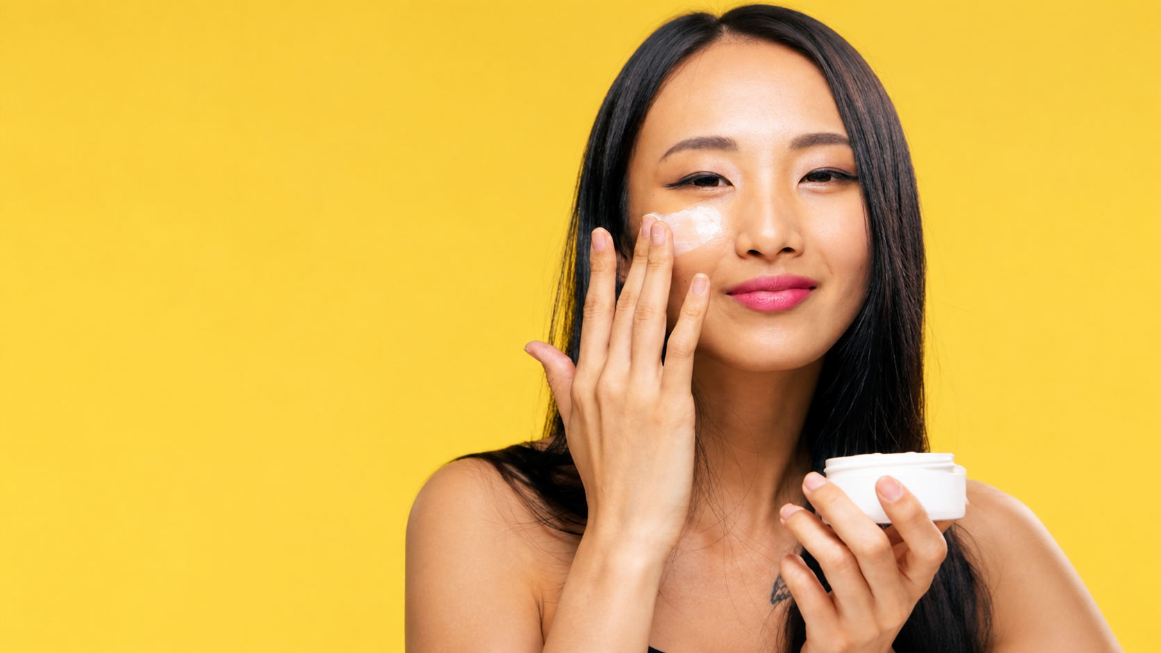 Woman applying white face cream to her cheek while holding an open cosmetic jar against a bright yellow background.