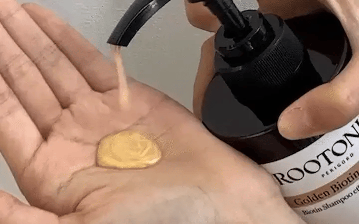 A close-up of a person's hand receiving a dollop of golden-yellow shampoo from a black pump bottle labeled ROOTONE Golden Biond Rinse.